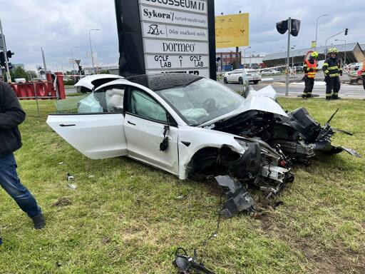 White Tesla with fully wrecked and burnt-out front section and deployed airbags, crashed backwards into Čestlice Shopping Center signpost after apparently having drifted across the grass. Some firefighters and onlookers visible.